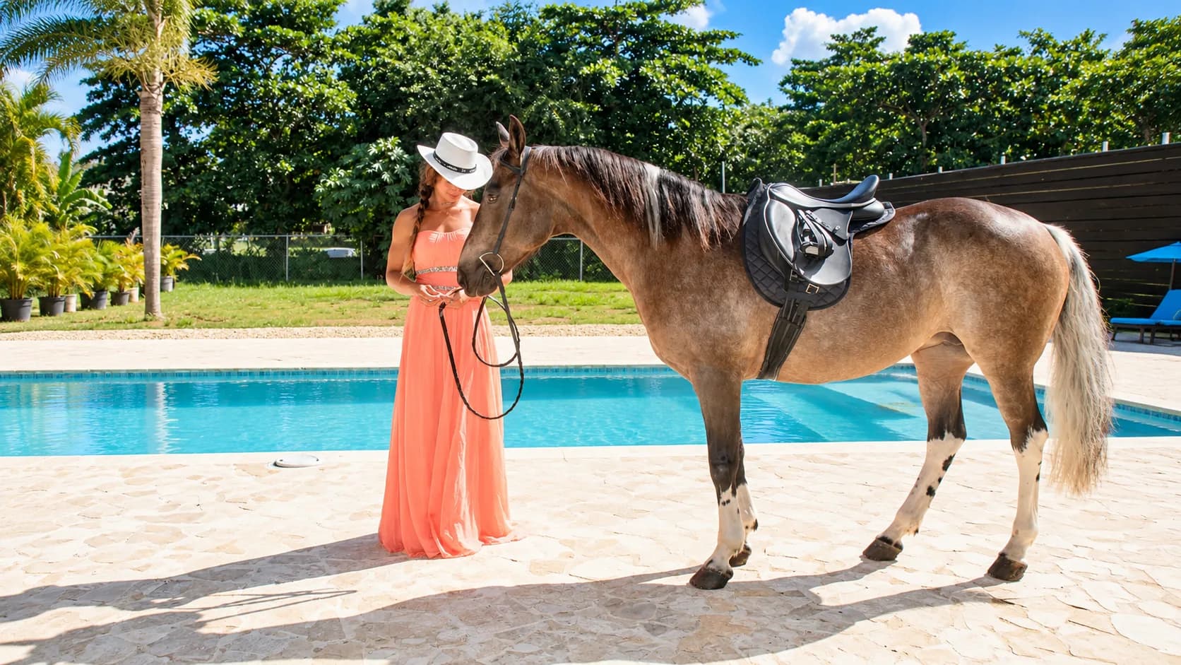 A woman in a long coral dress and a white hat holds the lead of a saddled light brown horse with white socks beside a swimming pool, with palm trees and green trees behind them.